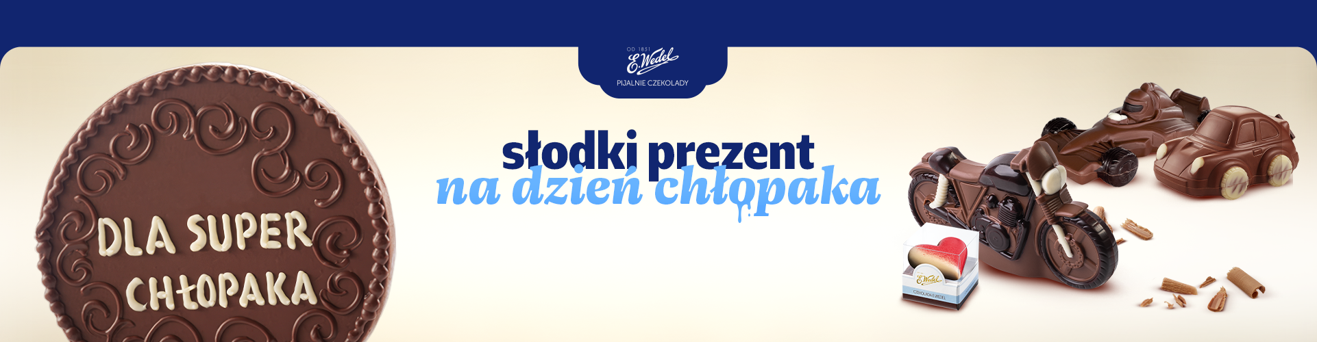 Grafika promująca prezenty na Dzień Chłopaka w Pijalniach Czekolady E.Wedel. Po lewej stronie znajduje się duża okrągła tabliczka czekolady z napisem „DLA SUPER CHŁOPAKA”. Na środku widnieje hasło „słodki prezent na dzień chłopaka”. Po prawej stronie pokazano czekoladowe figurki pojazdów: motocykl, bolid wyścigowy i samochód oraz małe opakowanie czekolady. Całość utrzymana jest w kremowo-granatowej kolorystyce z logo Pijalni Czekolady E.Wedel u góry.